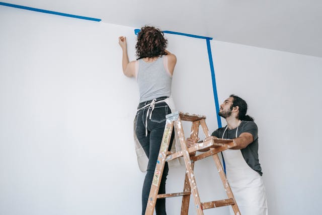 a woman on a step ladder wearing an apron as she removes painting tape from her ceiling. A man in an apron is holding the step ladder still.